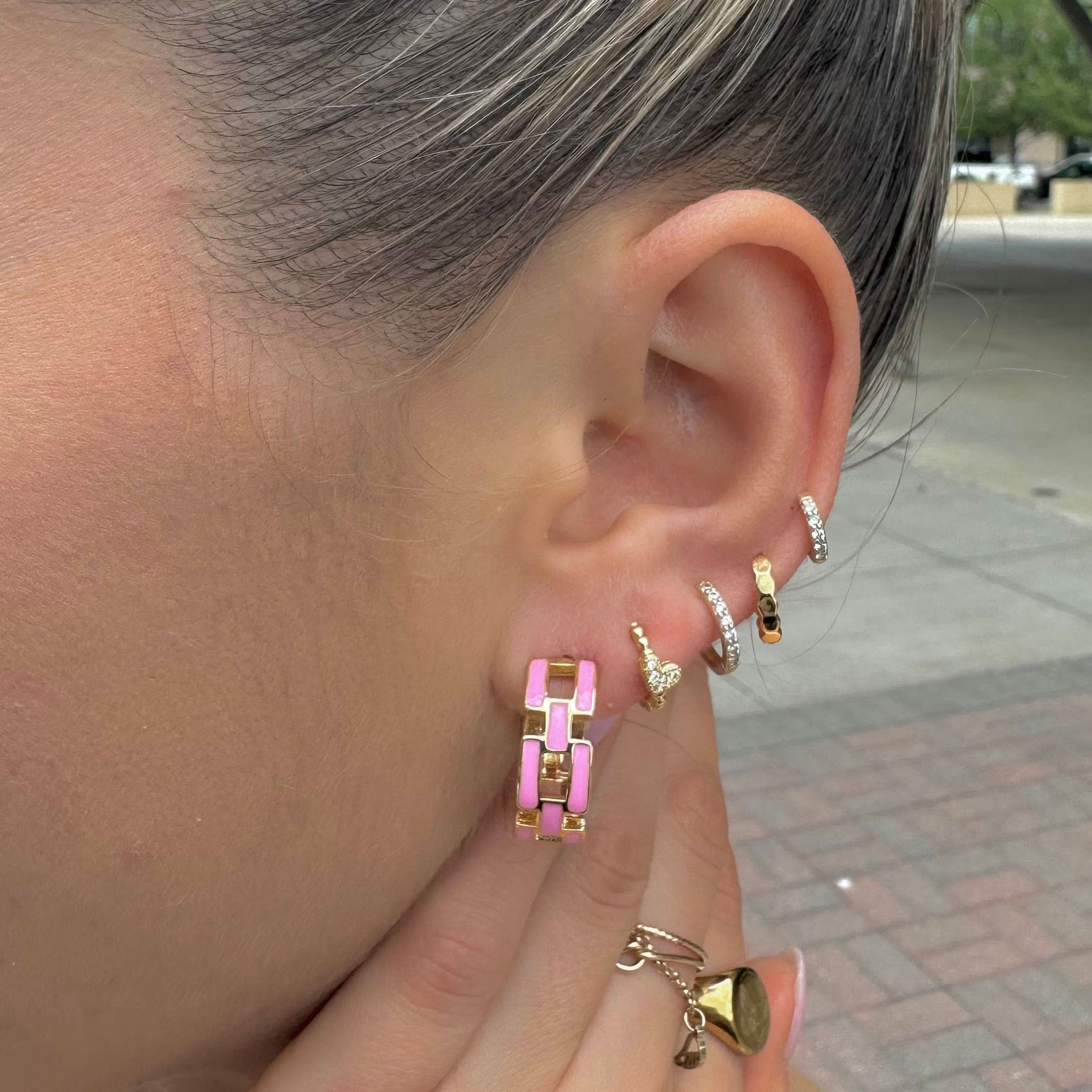 Close-up of an ear with multiple gold hoop earrings, including a pink one, with a blurred outdoor background.