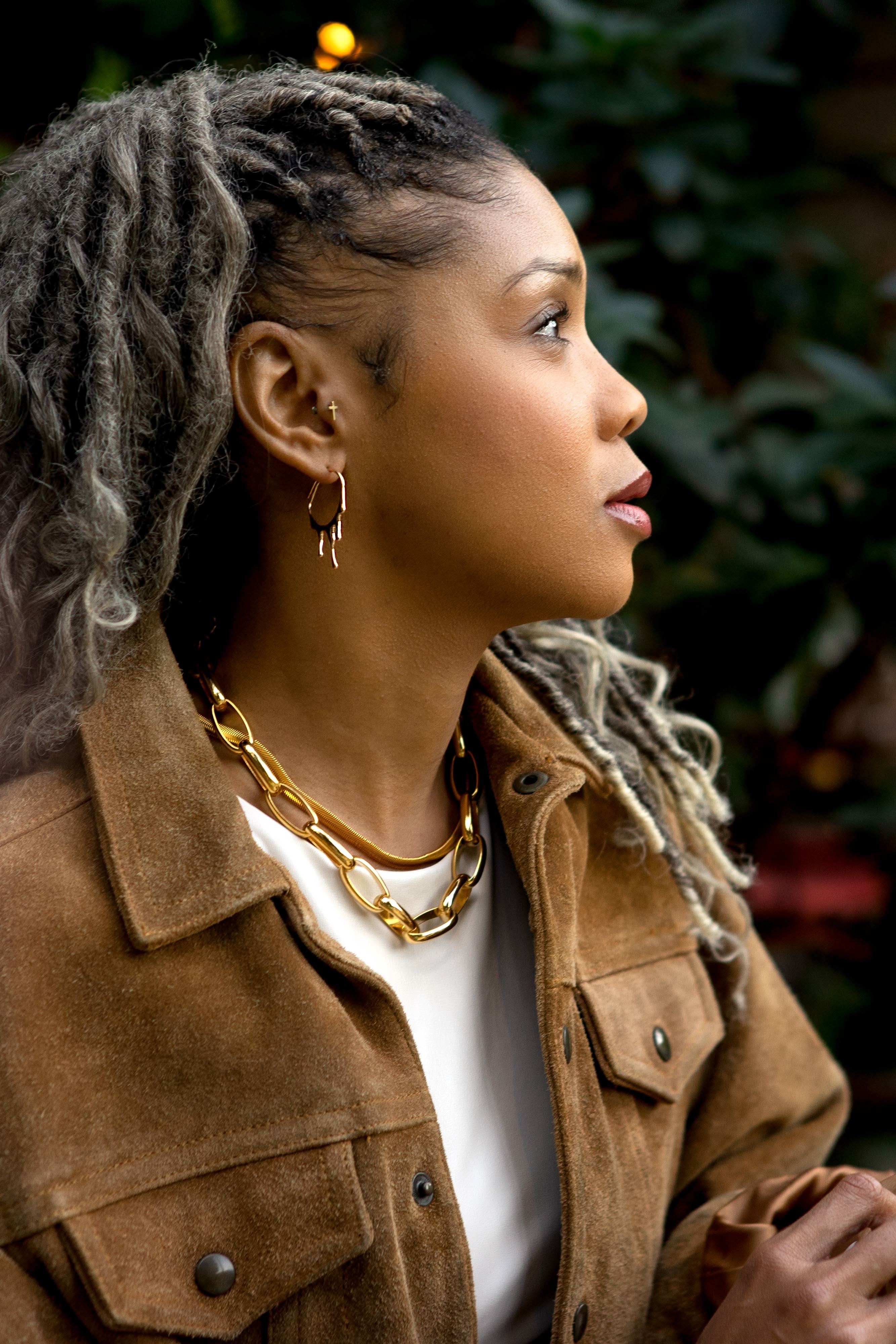 Woman wearing a brown jacket with a gold chain necklace, looking to the side.