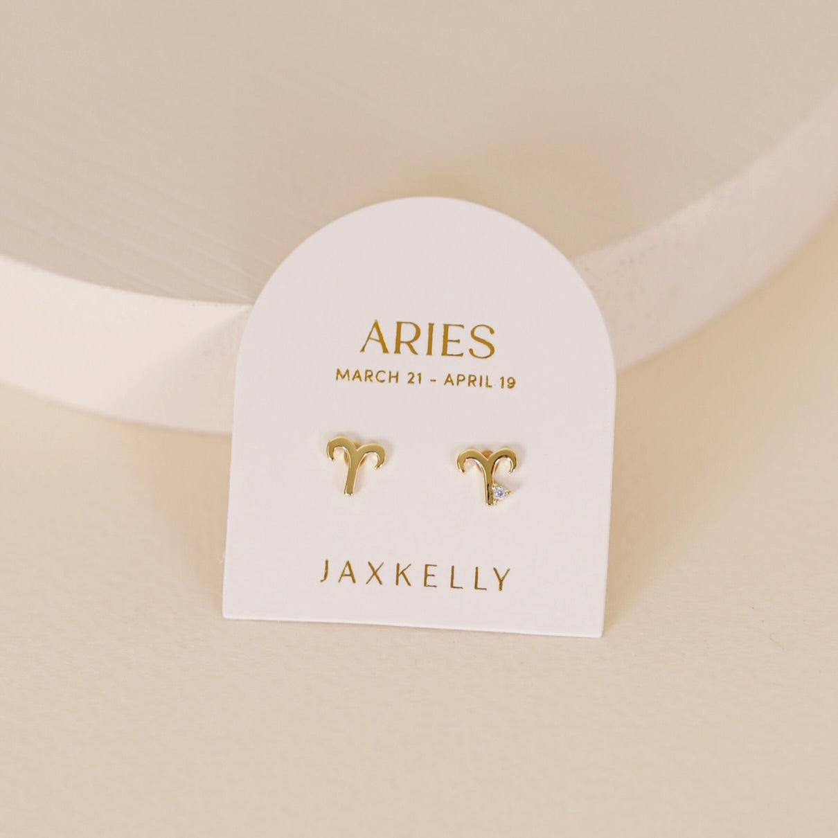 Gold zodiac charm studs in the shape of Aries astrological sign displayed on earring card