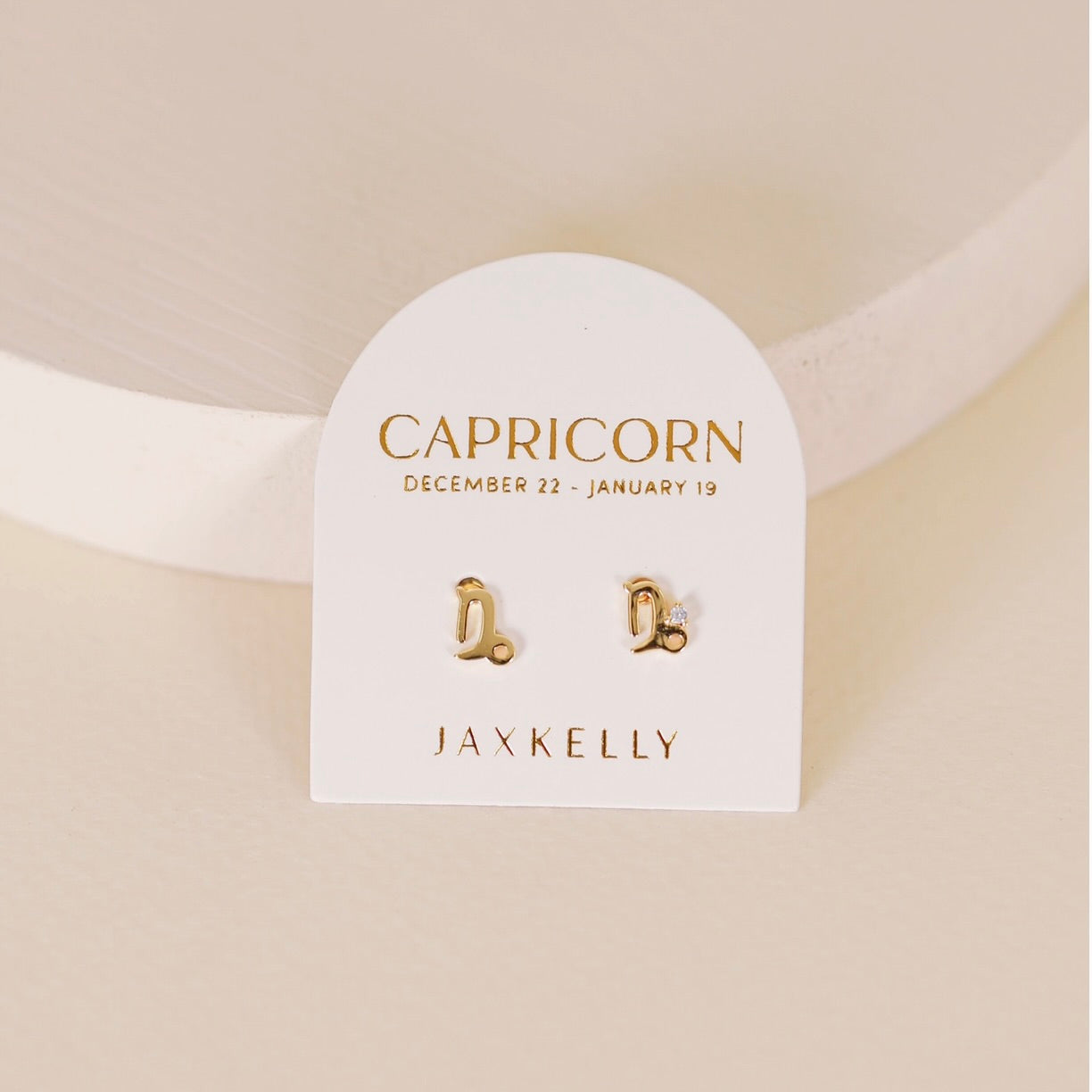 Gold zodiac charm studs in the shape of Capricorn astrological sign displayed on earring card