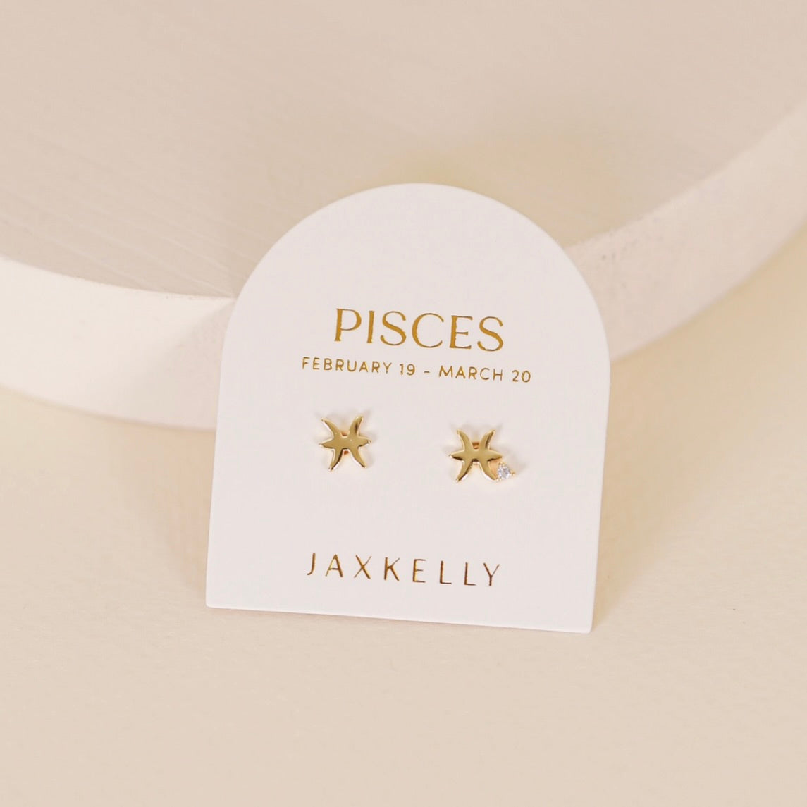 Gold zodiac charm studs in the shape of Pisces astrological sign displayed on earring card