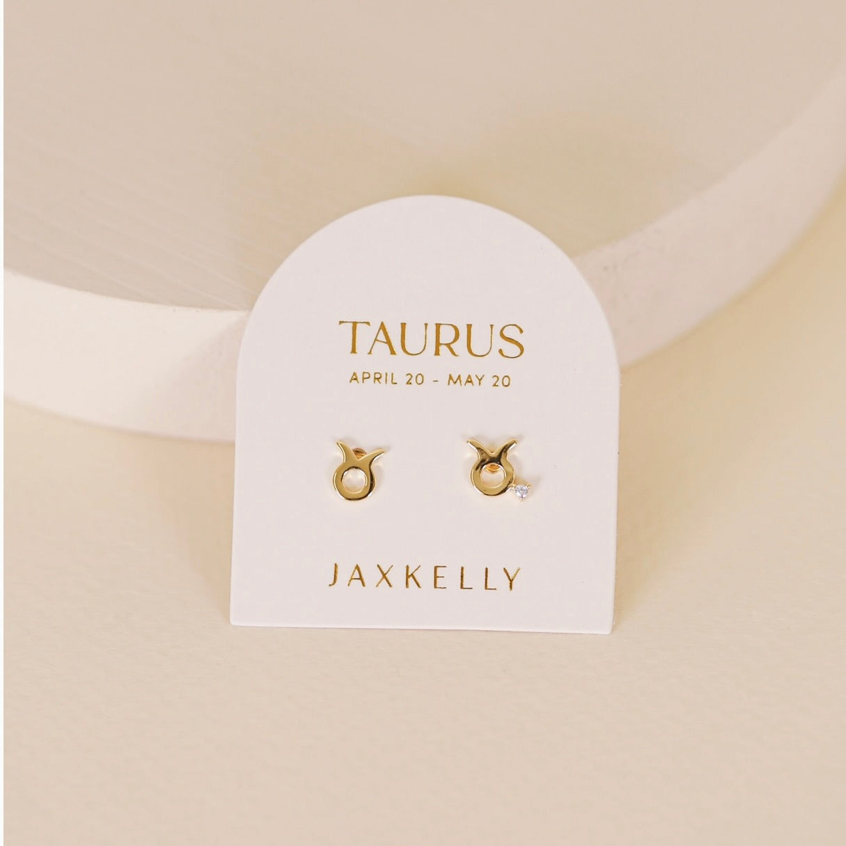 Gold zodiac charm studs in the shape of Taurus astrological sign displayed on earring card