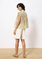 Woman turned to the side wearing flowing sleeveless pastel floral blouse with collar paired with white Bermuda shorts