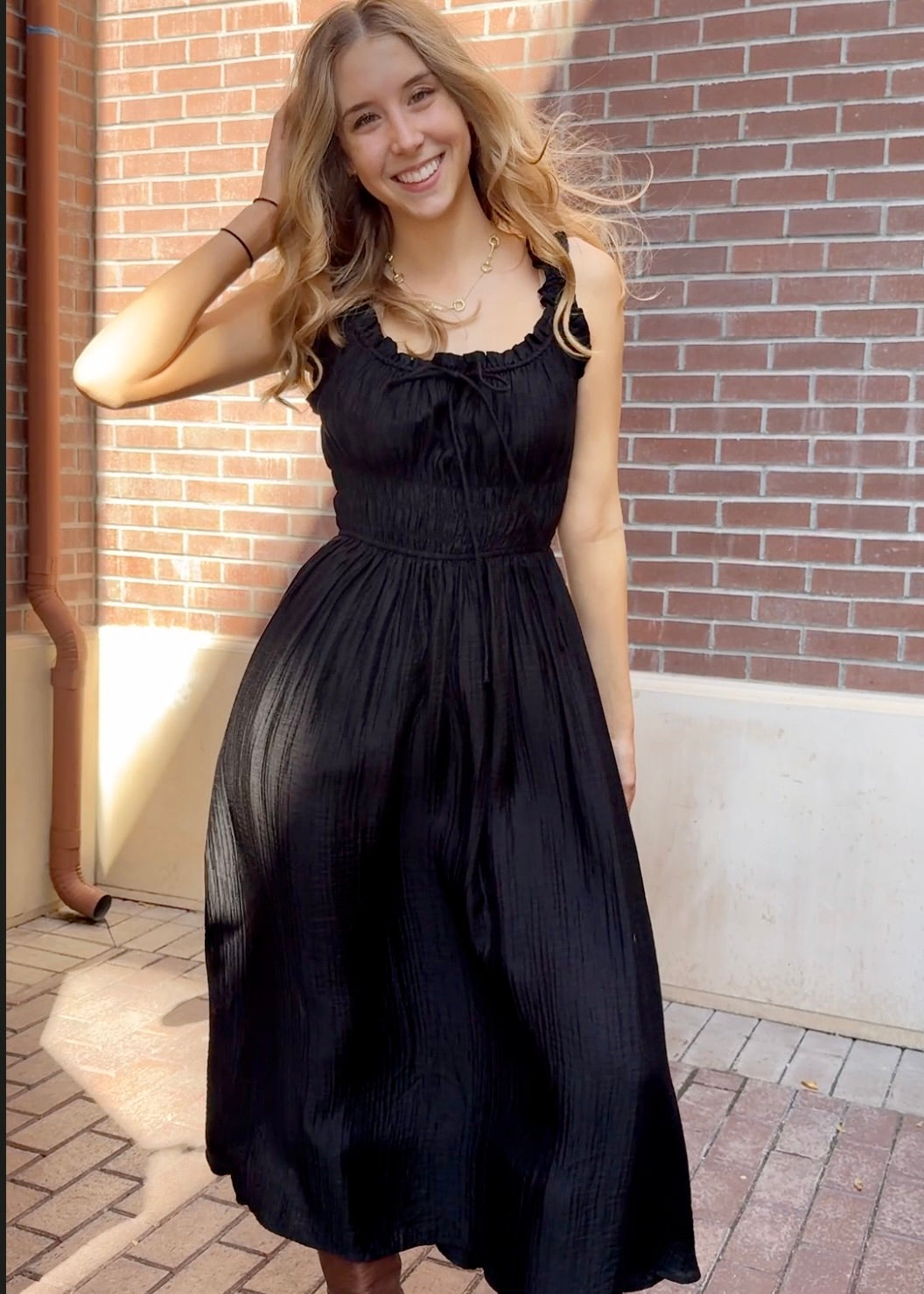 Model outside against a brick building wearing long black textured dress with thick straps 