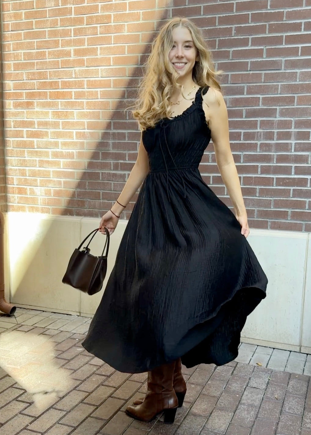 Model outside against a brick building wearing long black textured dress with thick straps, brown boots and brown purse