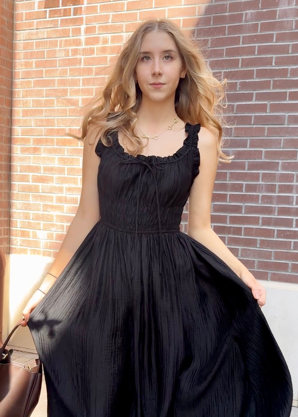 Model outside against a brick building wearing long black textured dress with thick straps holding brown purse