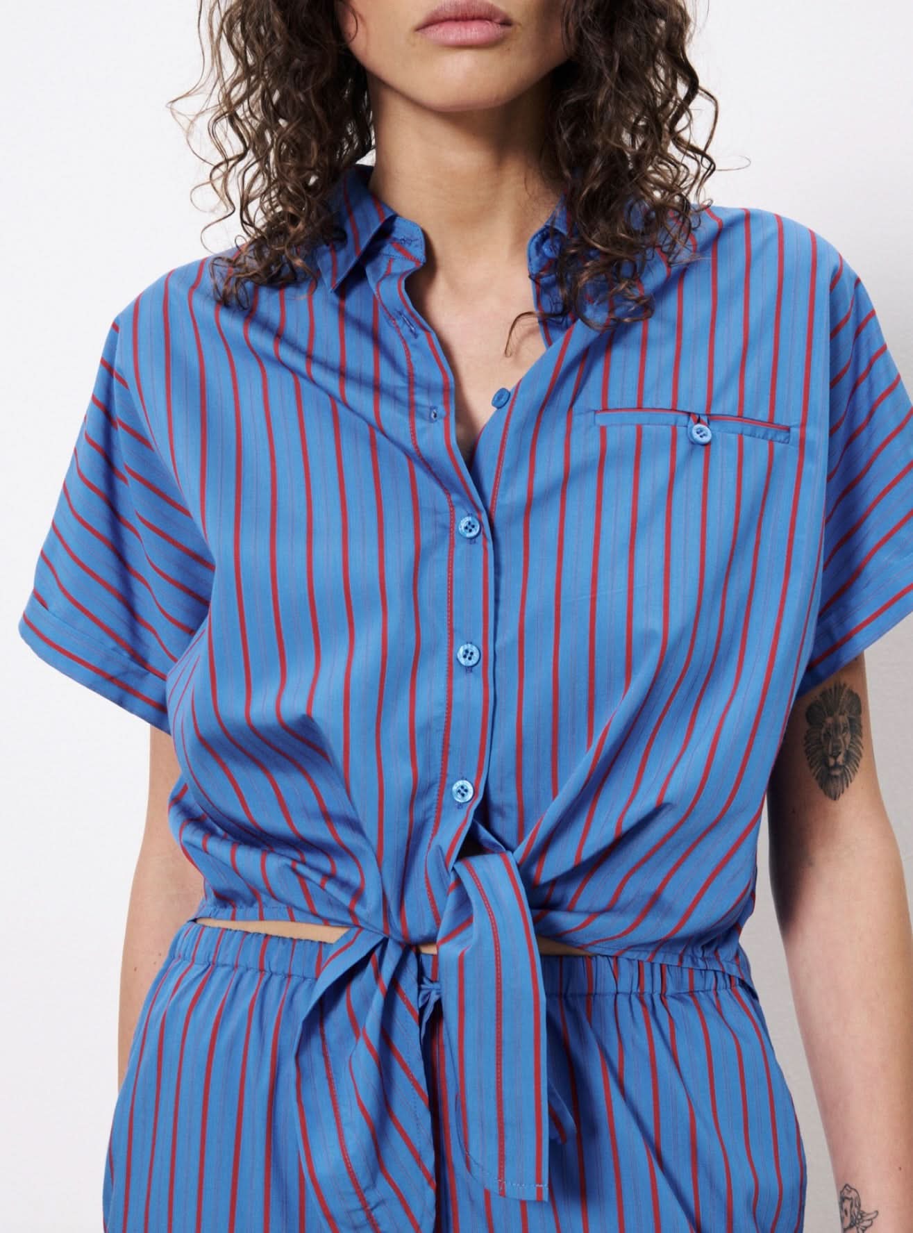 Woman wearing blue botton up blouse with vertical red stripes and a front pocket. Wearing pants with the same pattern.