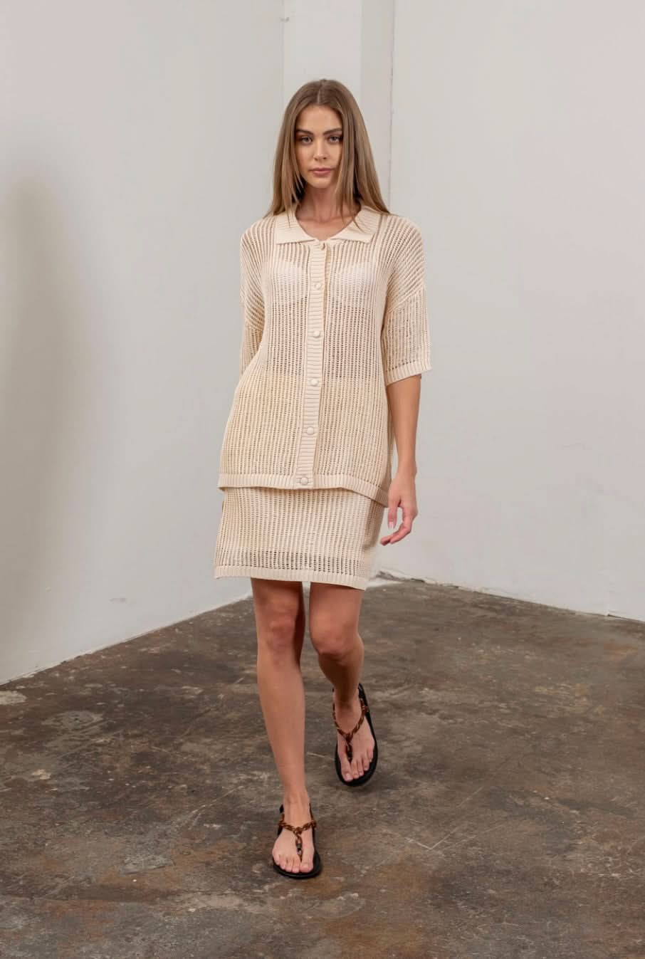 Woman walking toward the camera wearing crochet tan beige colored collared button up shirt with matching skirt 