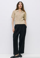 Woman wearing beige short-sleeve open knit top, collared neckline zip front closure paired black dress pants 