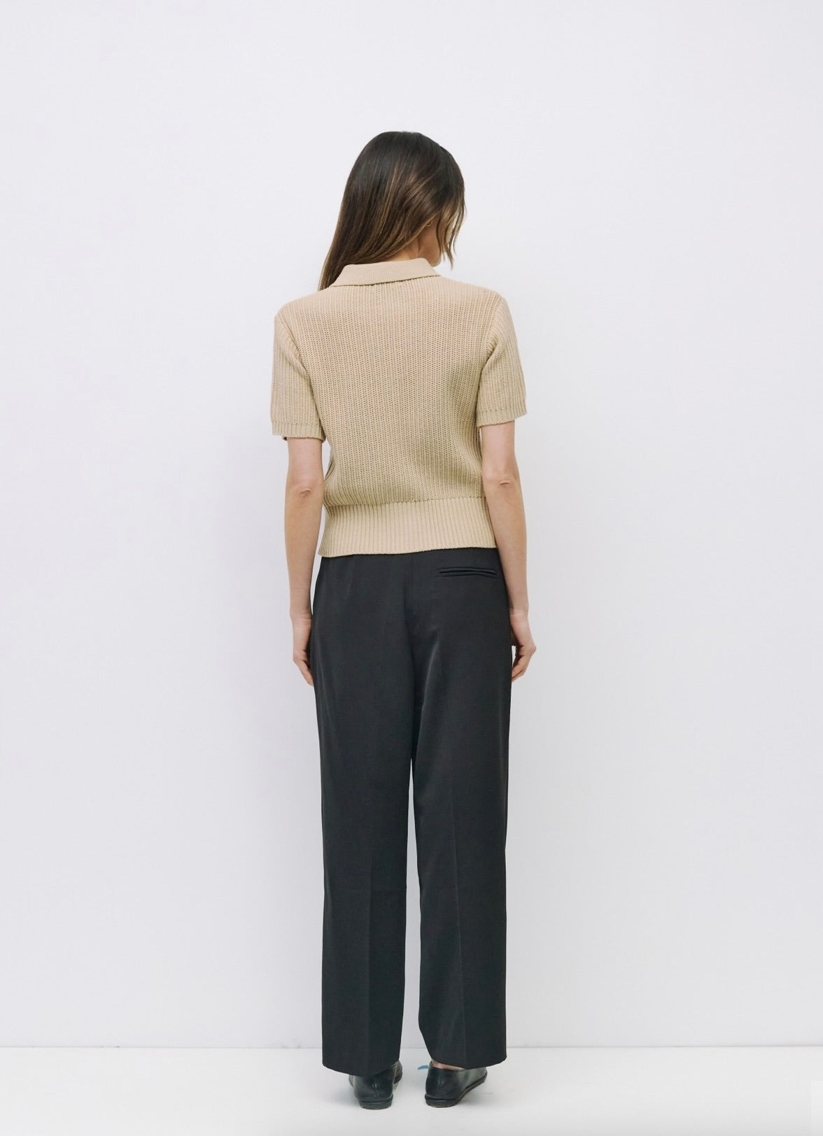 Woman facing backward wearing beige short-sleeve open knit top, collared neckline paired with black dress pants 