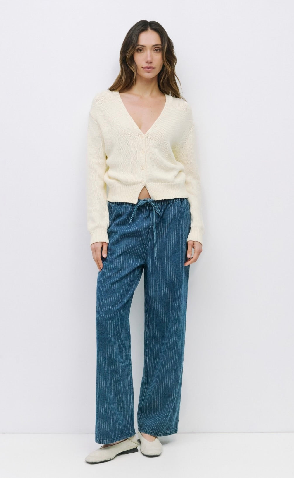 Woman wearing off white knit long sleeve sweater deep v neck with front button closure paired with pinstriped blue drawstring jeans 