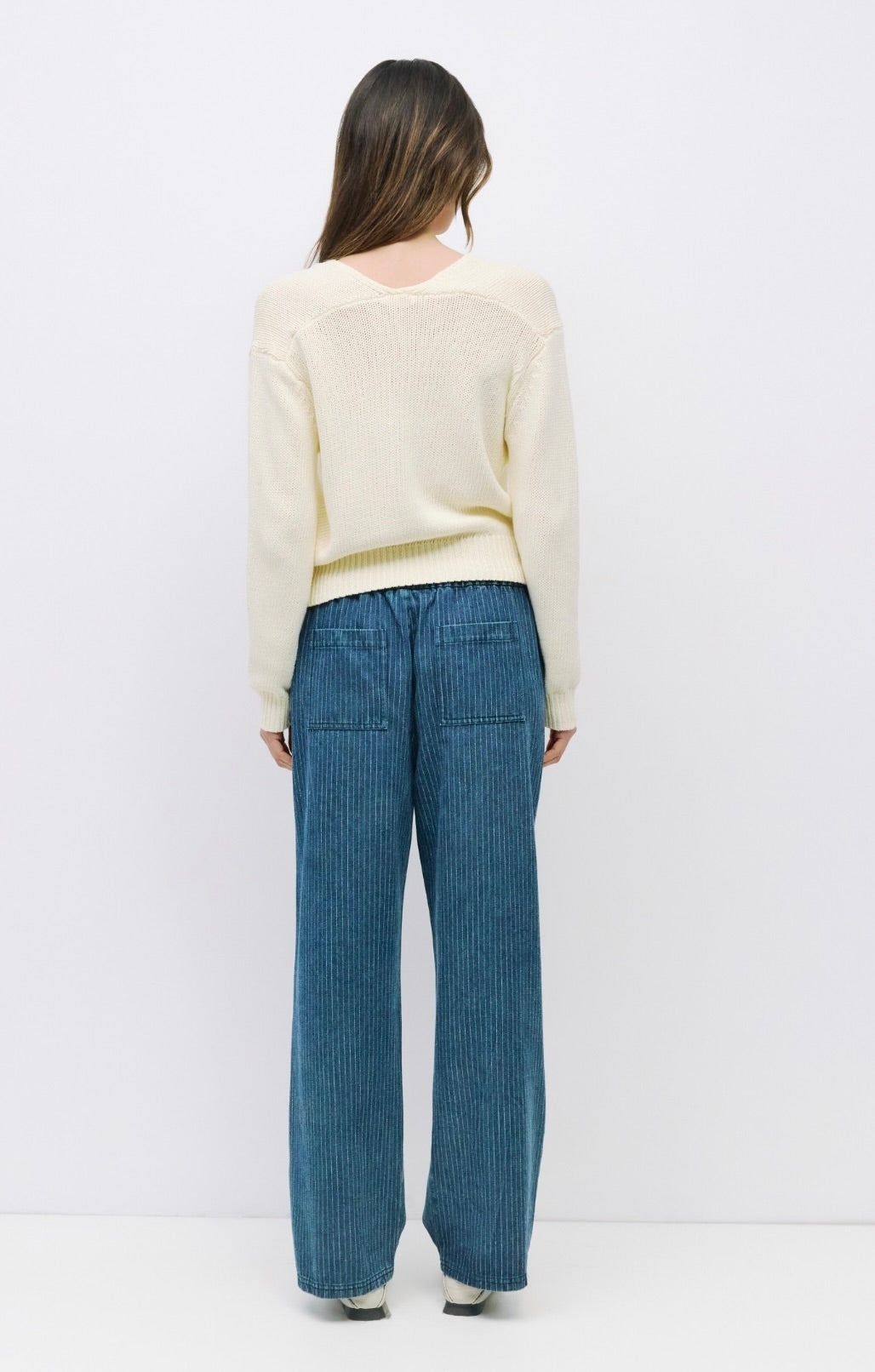 Woman facing backward wearing off white knit long sleeve sweater paired with pinstriped blue drawstring jeans 