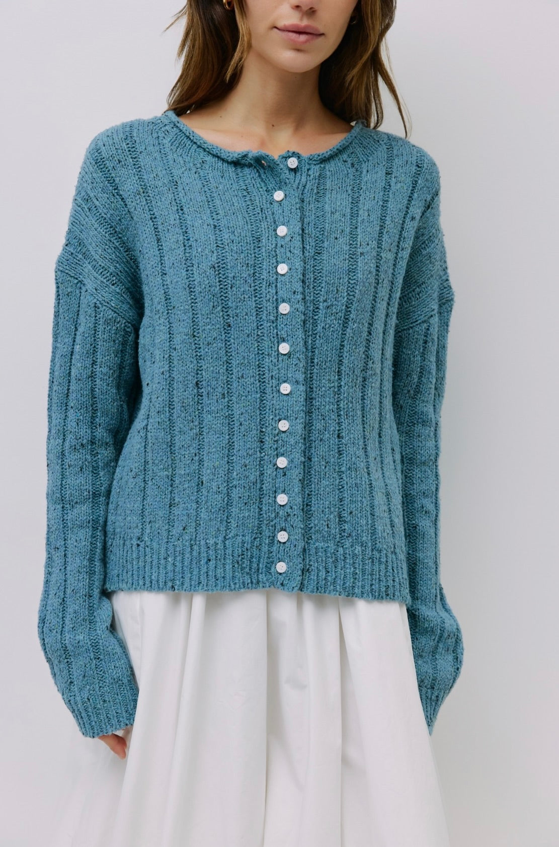 Woman wearing Blue specked chunky ribbed knit button down cardigan with rolled crewneck, dropped shoulders, relaxed fit and white skirt 