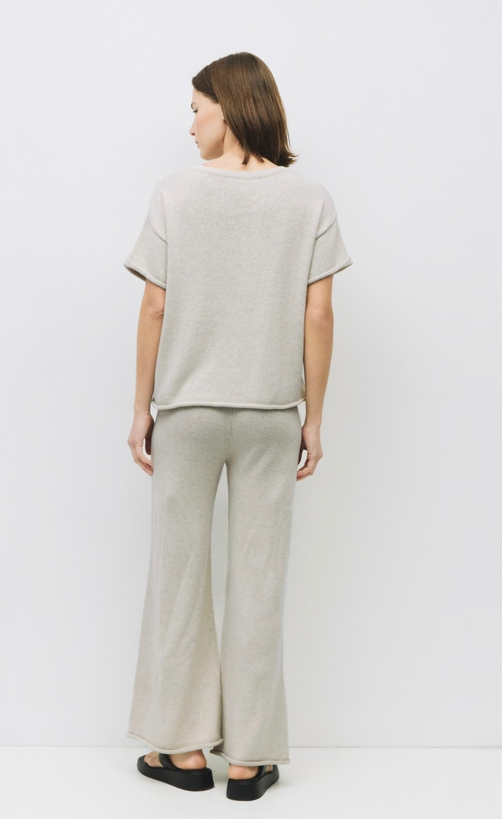 Woman wearing oatmeal color wide-leg knit trousers with a relaxed fit and raw roll-up hem and matching shirt facing backward 