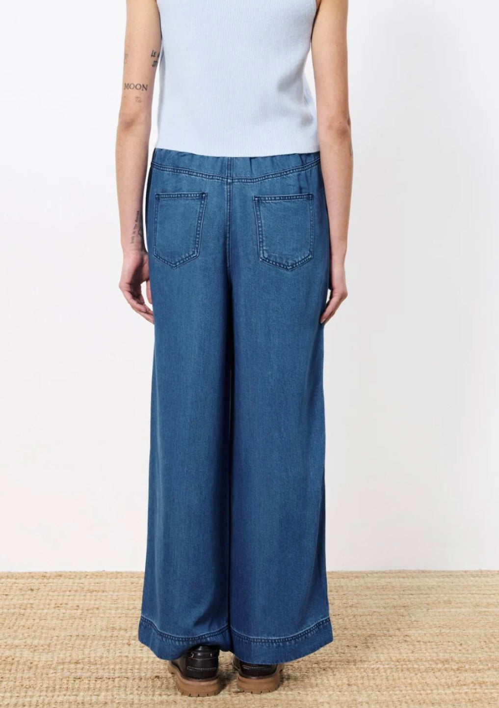 Woman facing backward wearing wide leg denim with elastic waistband  two back patch pockets paired with light blue top and black shoes 