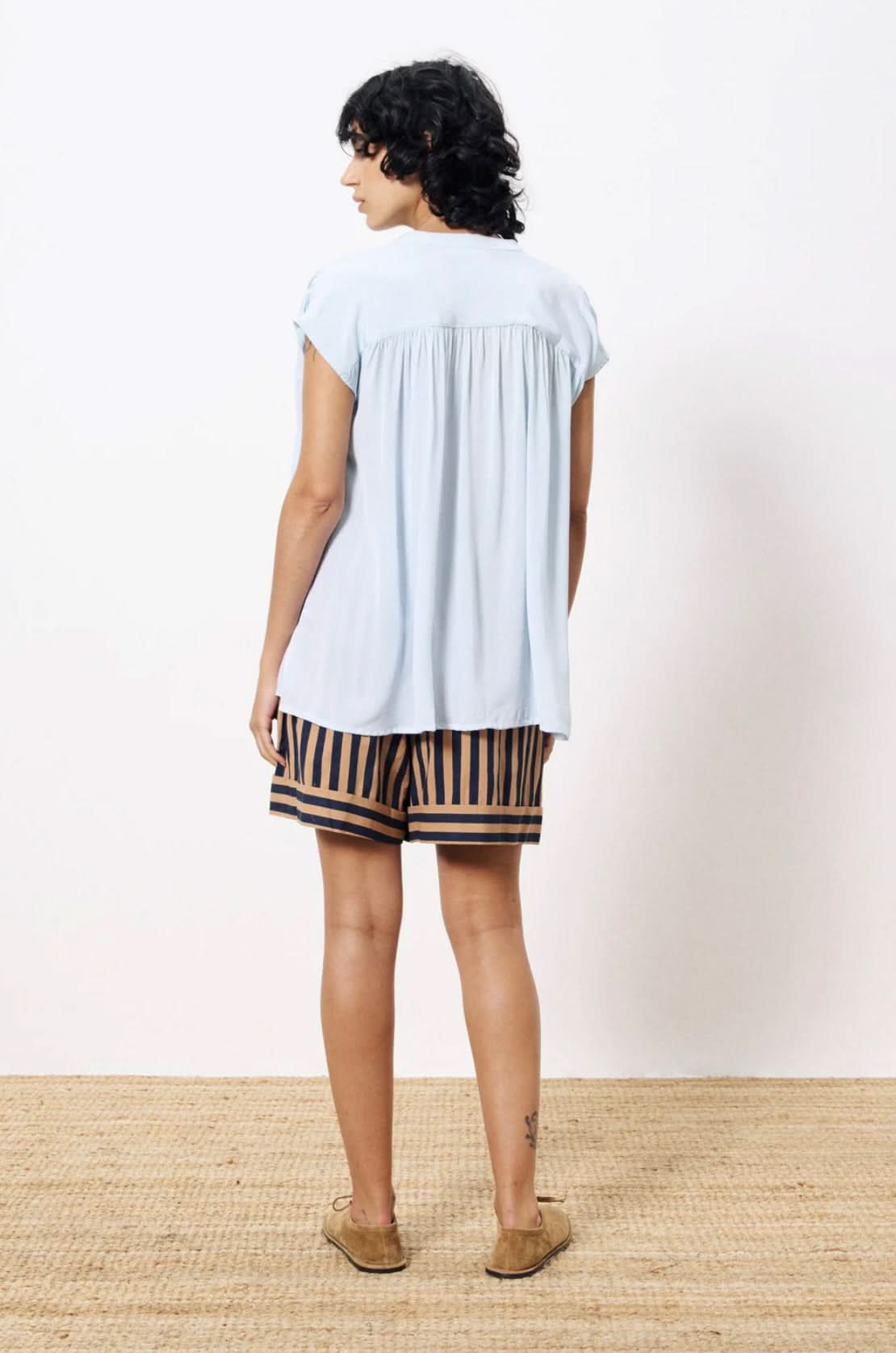 Woman turned backward wearing lightweight light blue blouse paired with tan and black dress shorts 