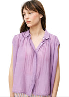 Woman wearing purple silky cotton button up, purple blouse, standing against white wall 