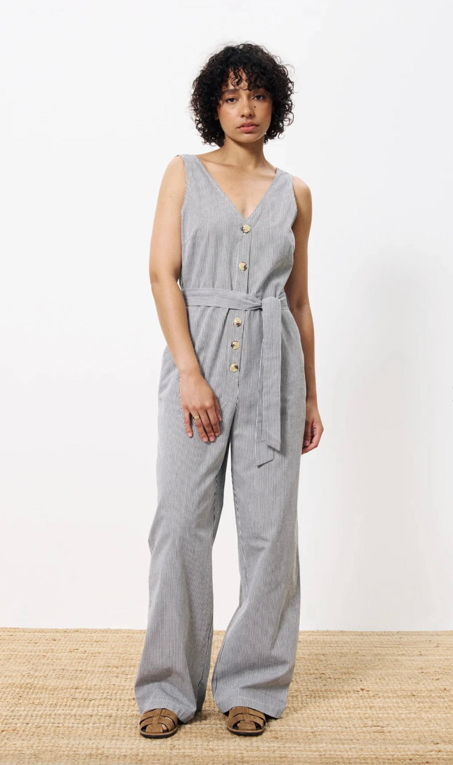 Woman wearing vertically striped grey and white jumpsuit with buttons down the front and a tie around waist 