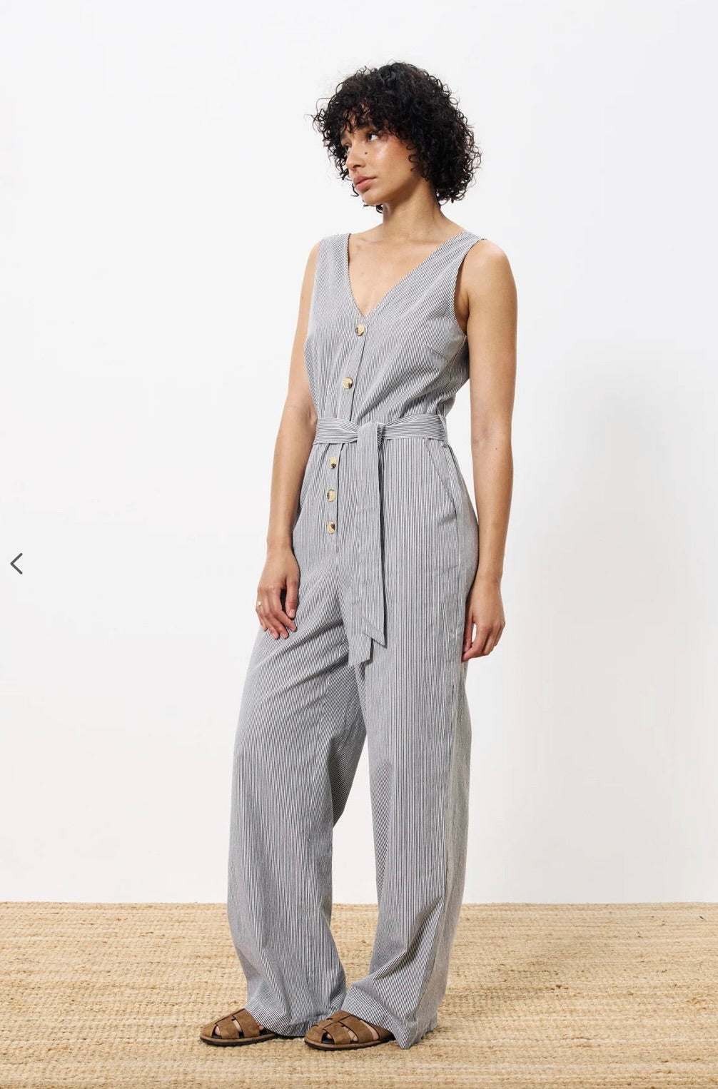 Woman wearing vertically pinstriped grey and white jumpsuit with buttons down the front and a tie around waist 