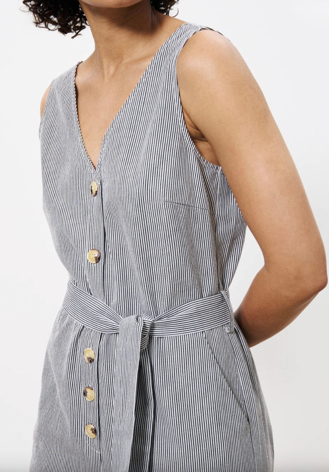 Woman wearing vertically pinstriped grey and white jumpsuit with buttons down the front and a tie around waist 
