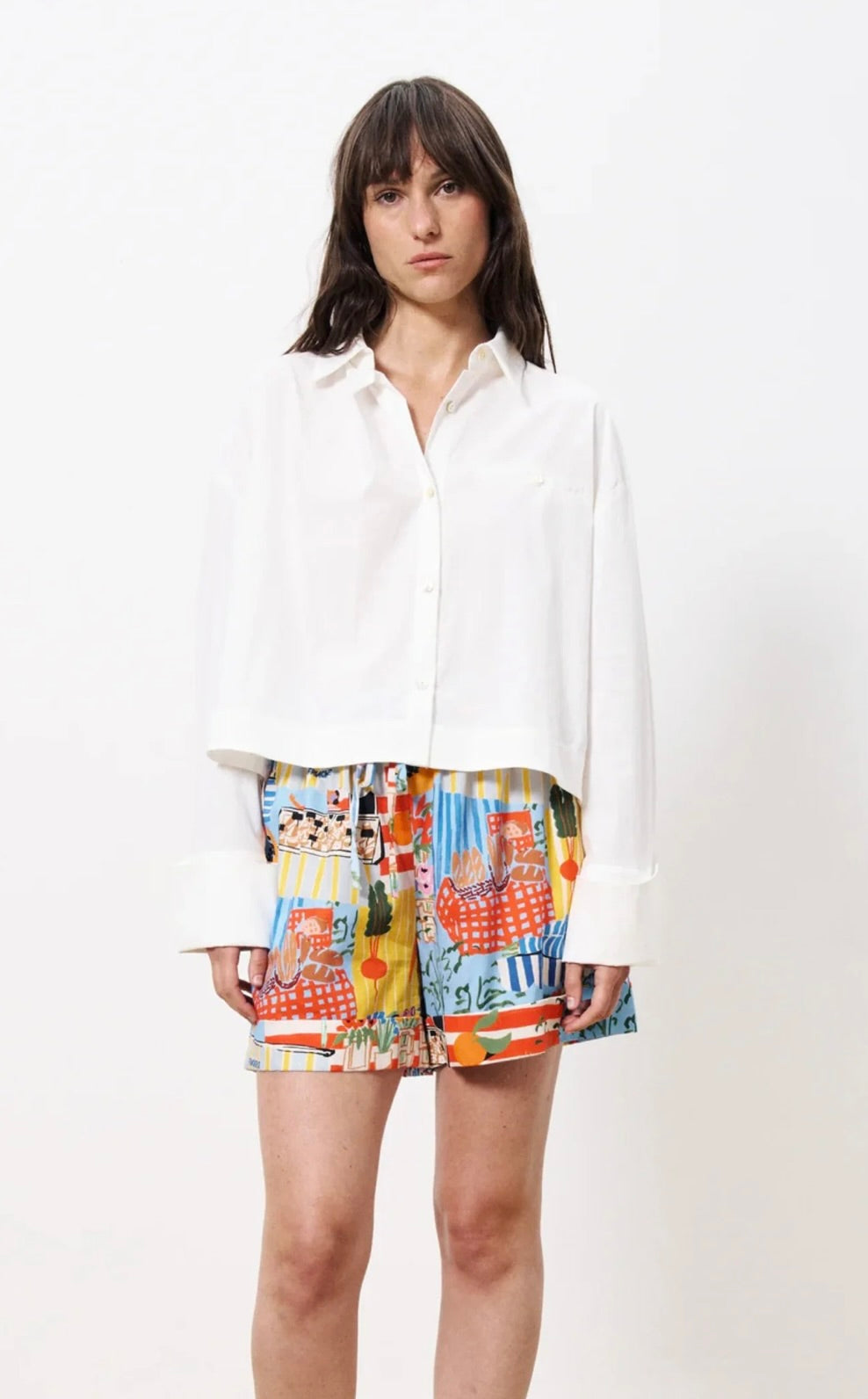 Woman wearing white shirt and unique design shorts depicting french country farmers market against a blue background with bread, fruit, vegetables, and many other items 