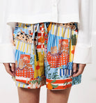 Woman wearing white shirt and unique design shorts depicting french country farmers market against a blue background with bread, fruit, vegetables, and many other items 