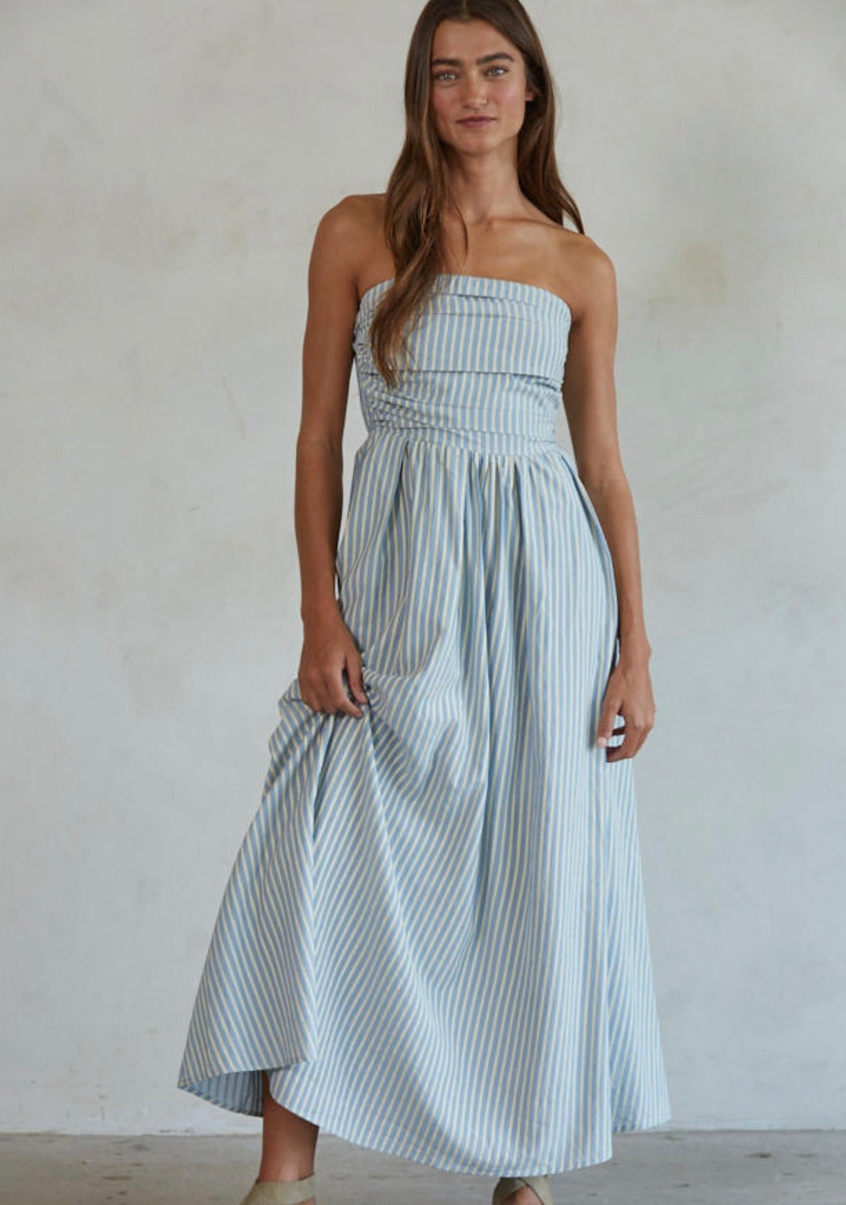 Woman wearing white and green/blue vertically striped, sleeveless maxi dress standing against a blank wall