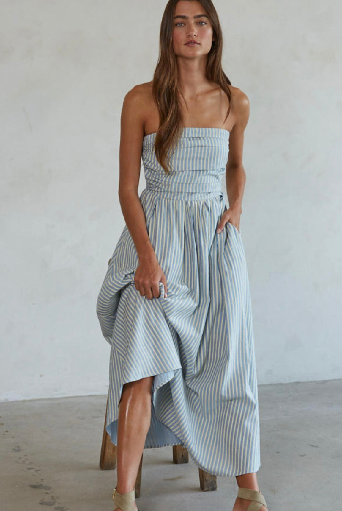 Woman wearing white and green/blue vertically striped, sleeveless maxi dress standing against a blank wall