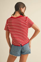 Woman wearing short sleeve red and taupe knitted sweater with jean shorts tuned backward to show 2 button closure on bottom of sweater 