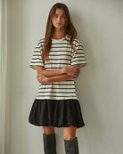 Woman crossing her arms wearing a short sleeve black and white striped thigh length dress with a black skirt behind a white background. 