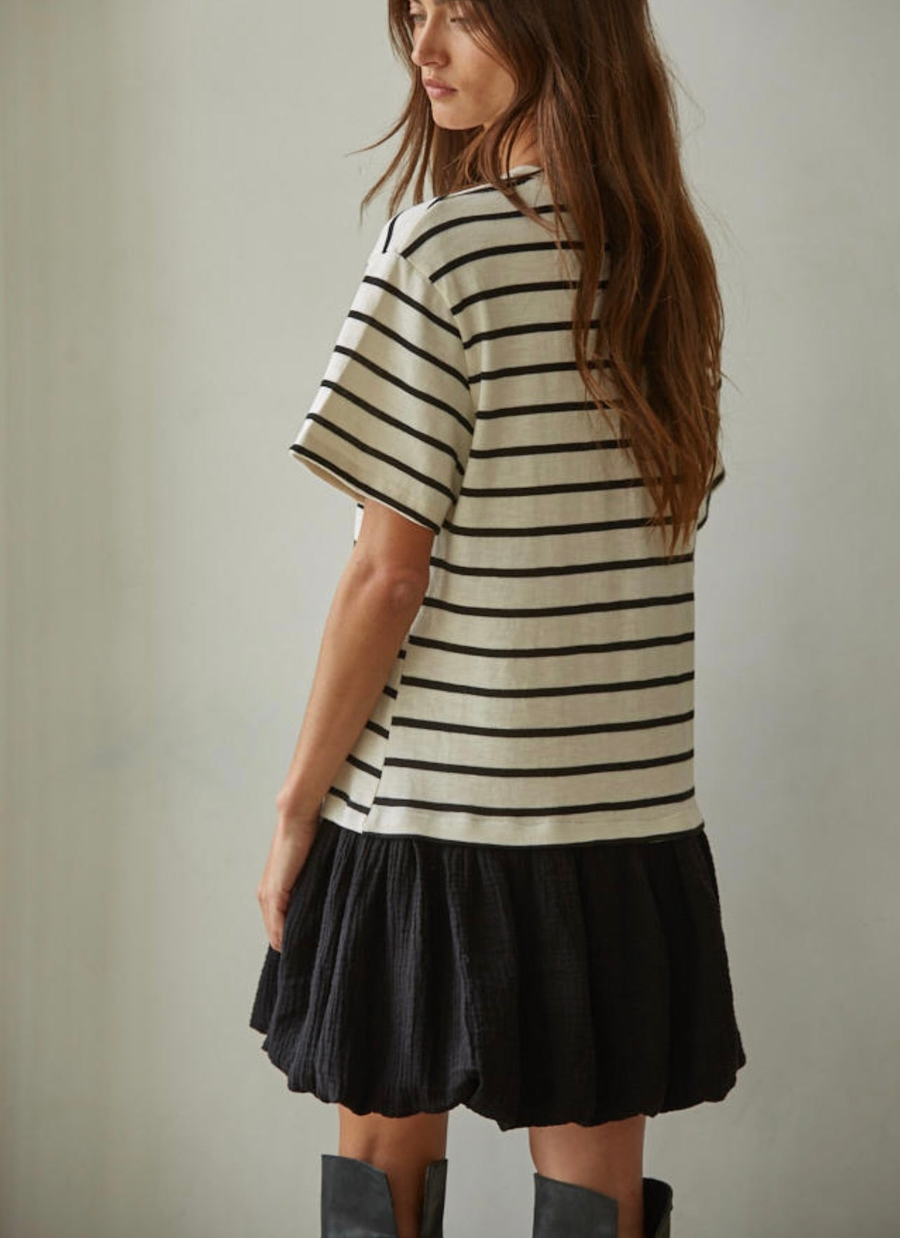 Woman with her back turned wearing a short sleeve black and white striped thigh length dress with a black skirt and black boots behind a white background. 