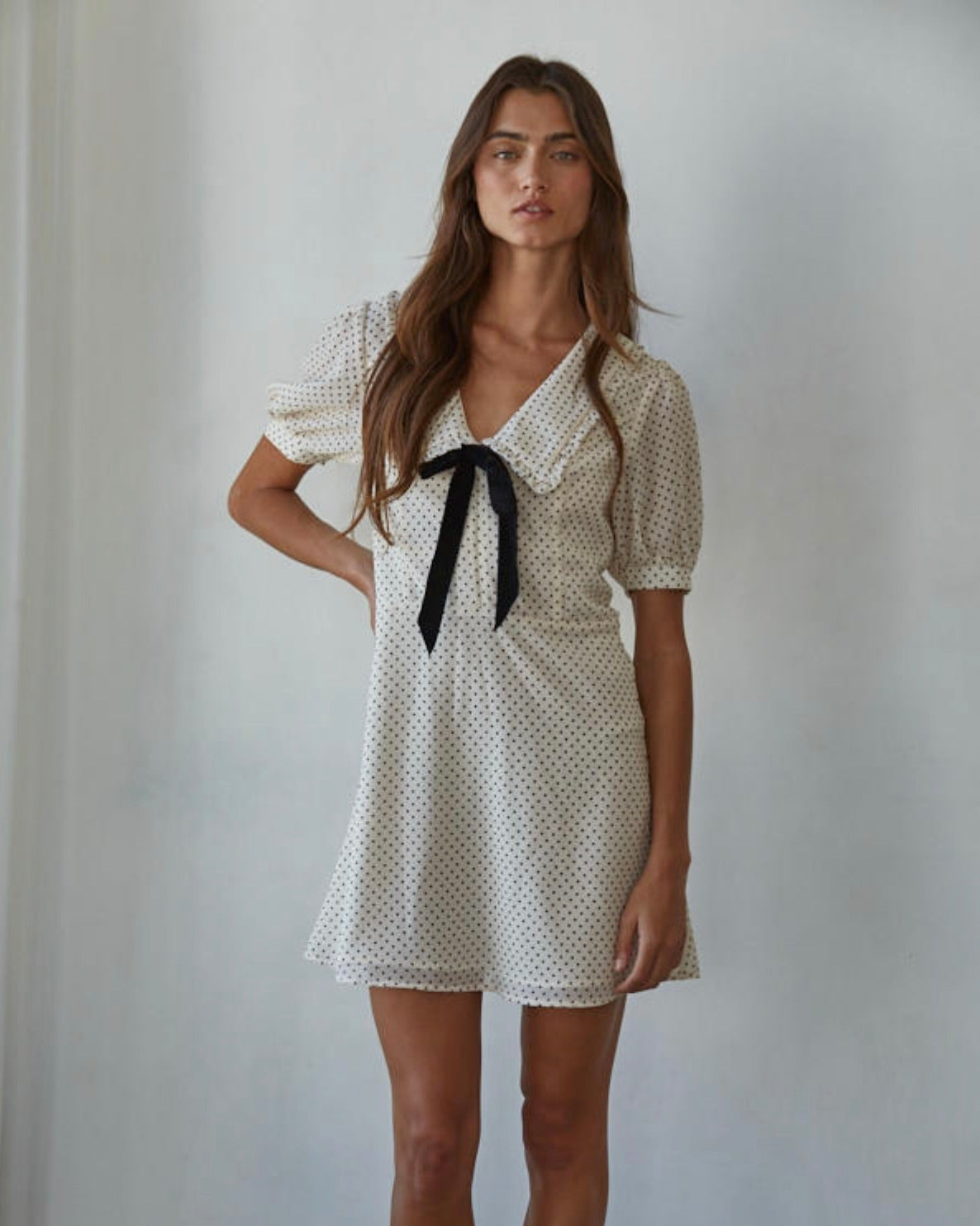 Woman wearing a thigh length polka dot dress with puffy sleeves and a black ribbon behind a white background. 