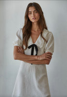 Woman wearing a thigh length polka dot dress with puffy sleeves and a black ribbon behind a white background. 