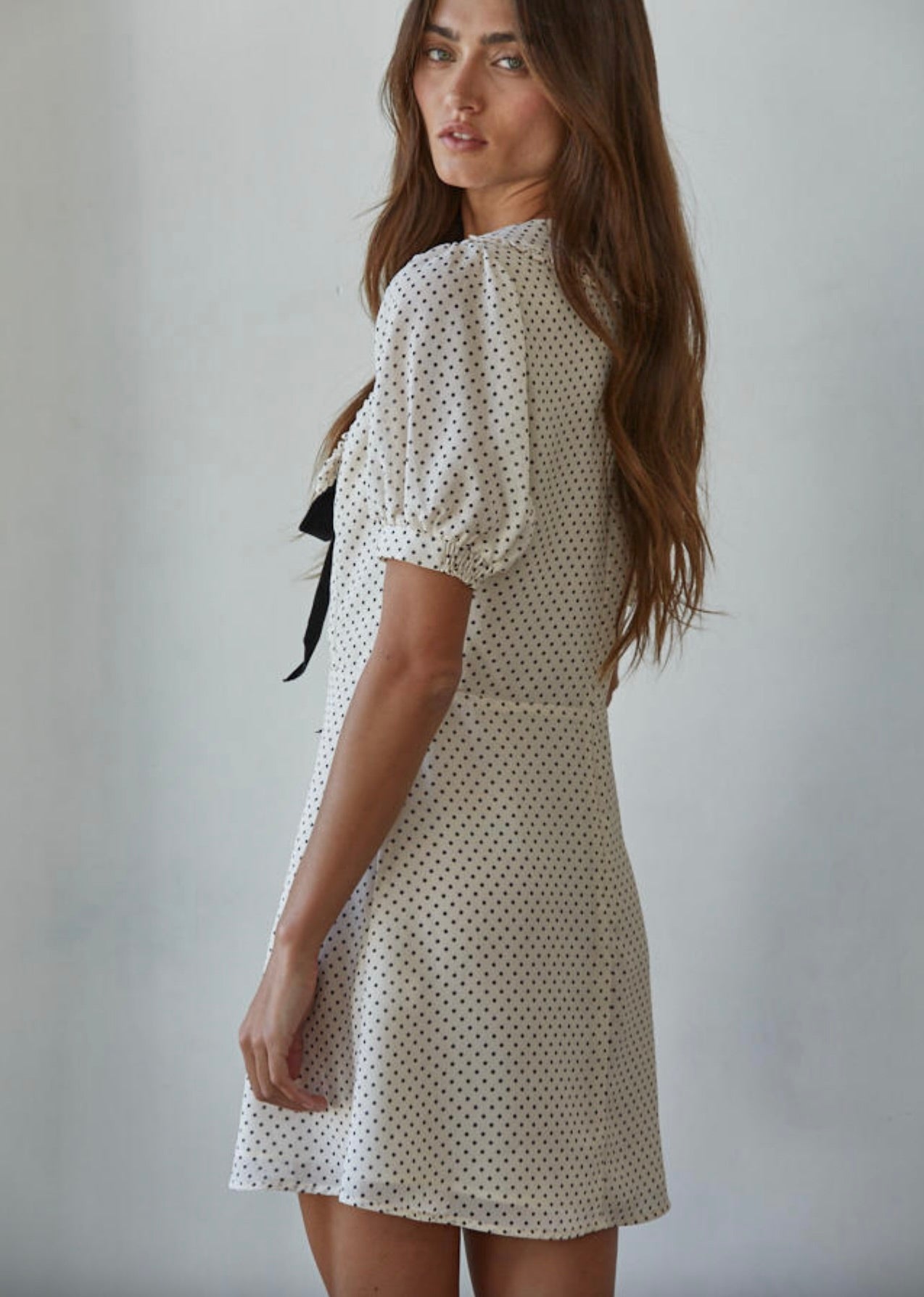 Woman wearing a thigh length polka dot dress with puffy sleeves and a black ribbon behind a white background. 