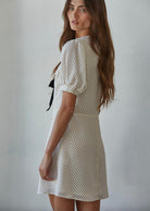 Woman wearing a thigh length polka dot dress with puffy sleeves and a black ribbon behind a white background. 