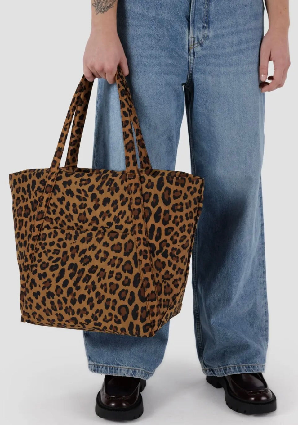 Man wearing jeans while holding a large leopard tote bag behind a white background. 
