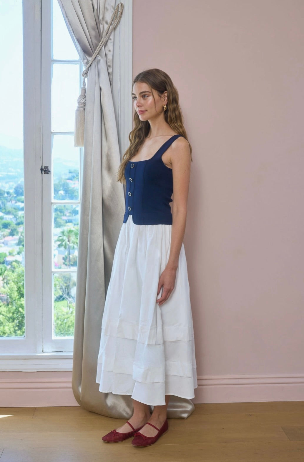 Woman wearing a blue top and white skirt standing in a room with large windows.