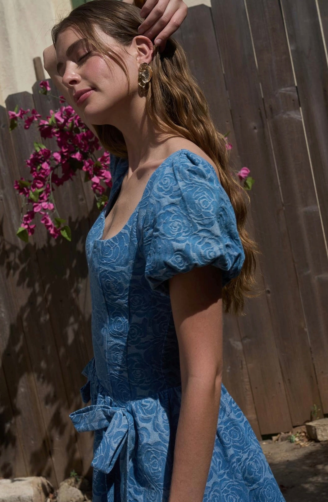 Woman in a blue lace dress standing outdoors with a wooden fence and flowers in the background