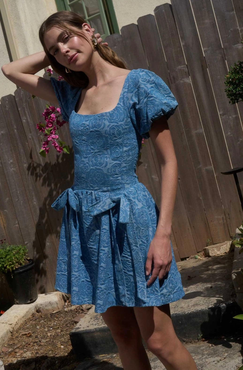 Woman wearing a blue dress standing outdoors with a wooden fence and plants in the background