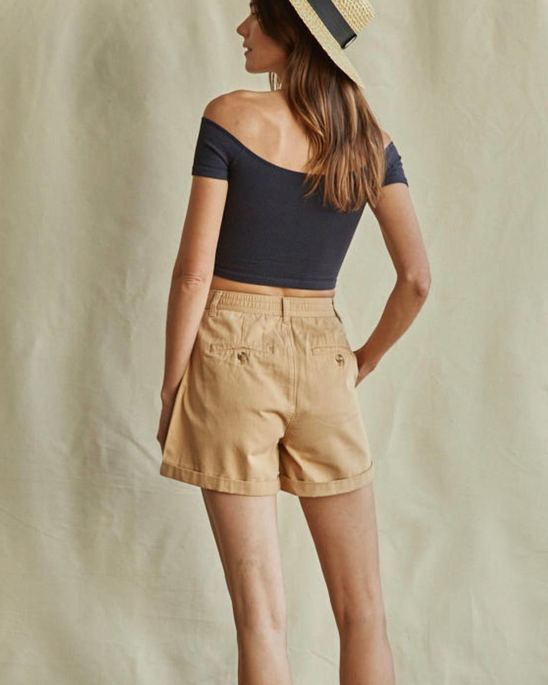 Woman wearing a navy top and beige shorts with a straw hat on a neutral background