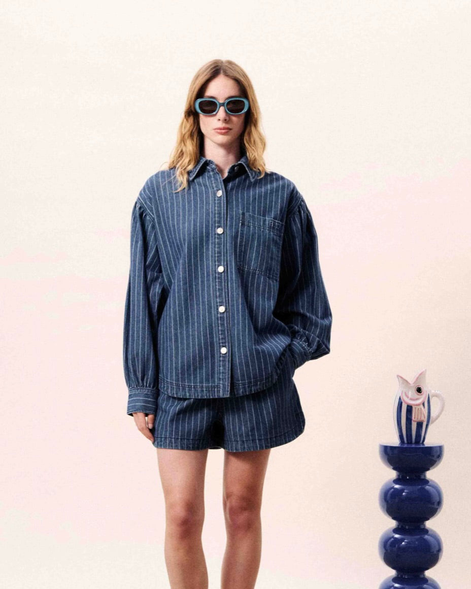 Woman wearing a blue striped shirt and shorts with sunglasses, standing against a light pink background.
