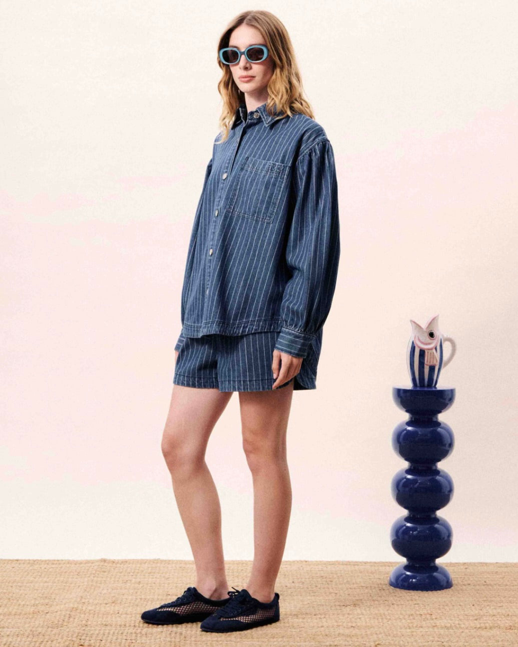 Woman wearing a blue striped outfit standing next to a decorative vase on a beige surface.