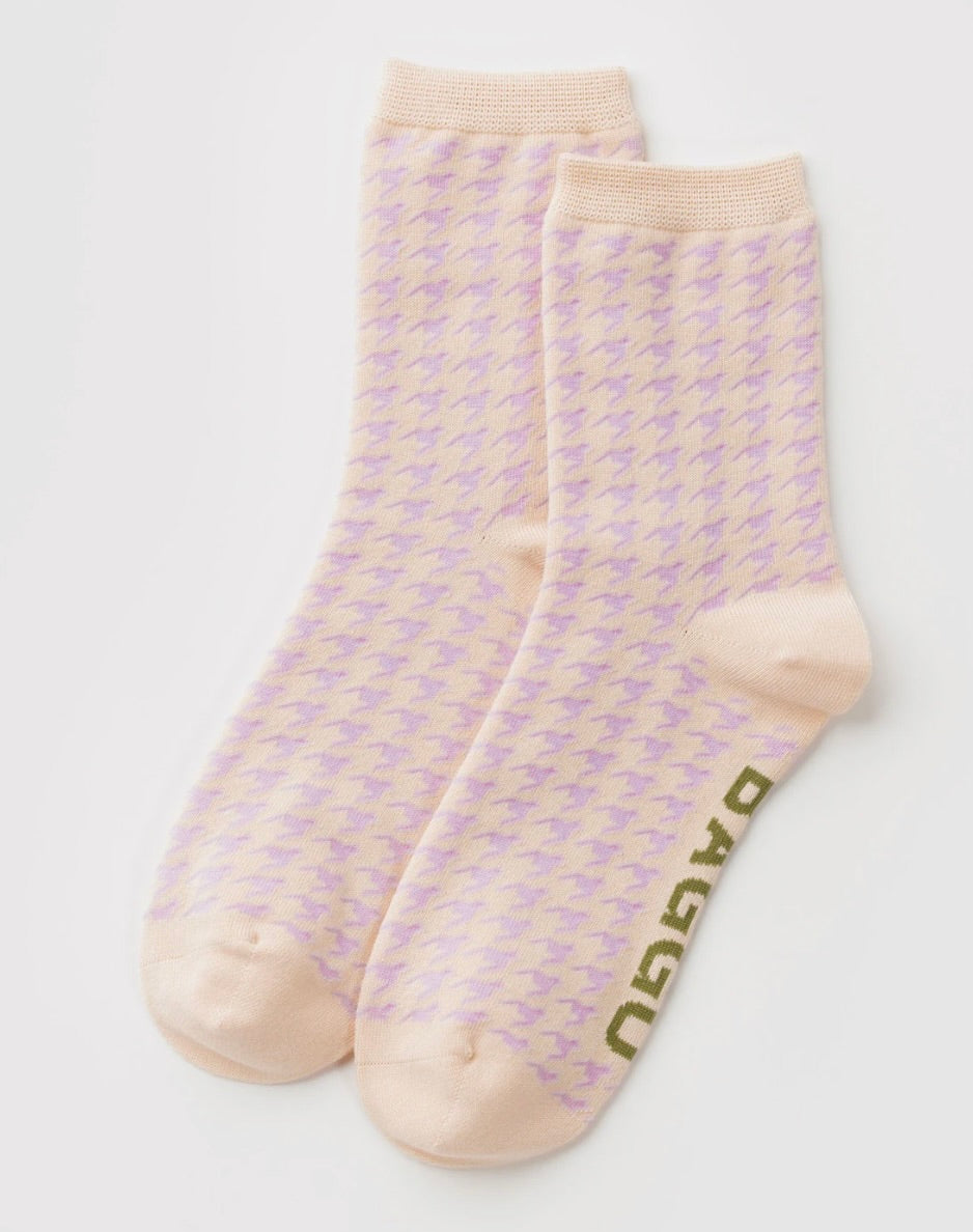 A pair of light peach crew socks with a repeating lavender houndstooth pattern and olive green 'DAGGU' branding printed on the sole, displayed on a clean white background.