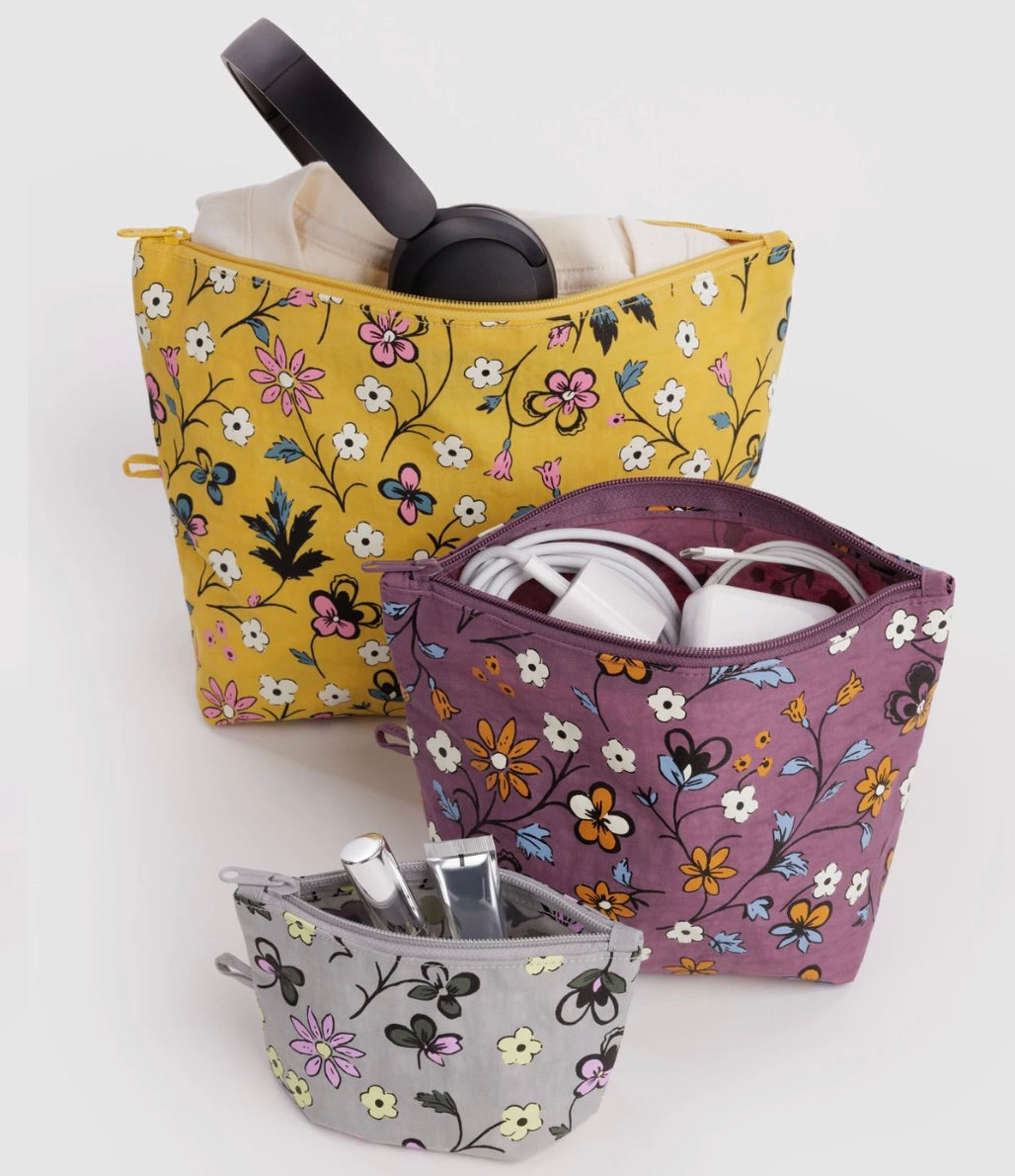 Three floral zippered cosmetic pouches in yellow, purple, and gray, shown with headphones, earbuds, and toiletries inside on a light gray background.