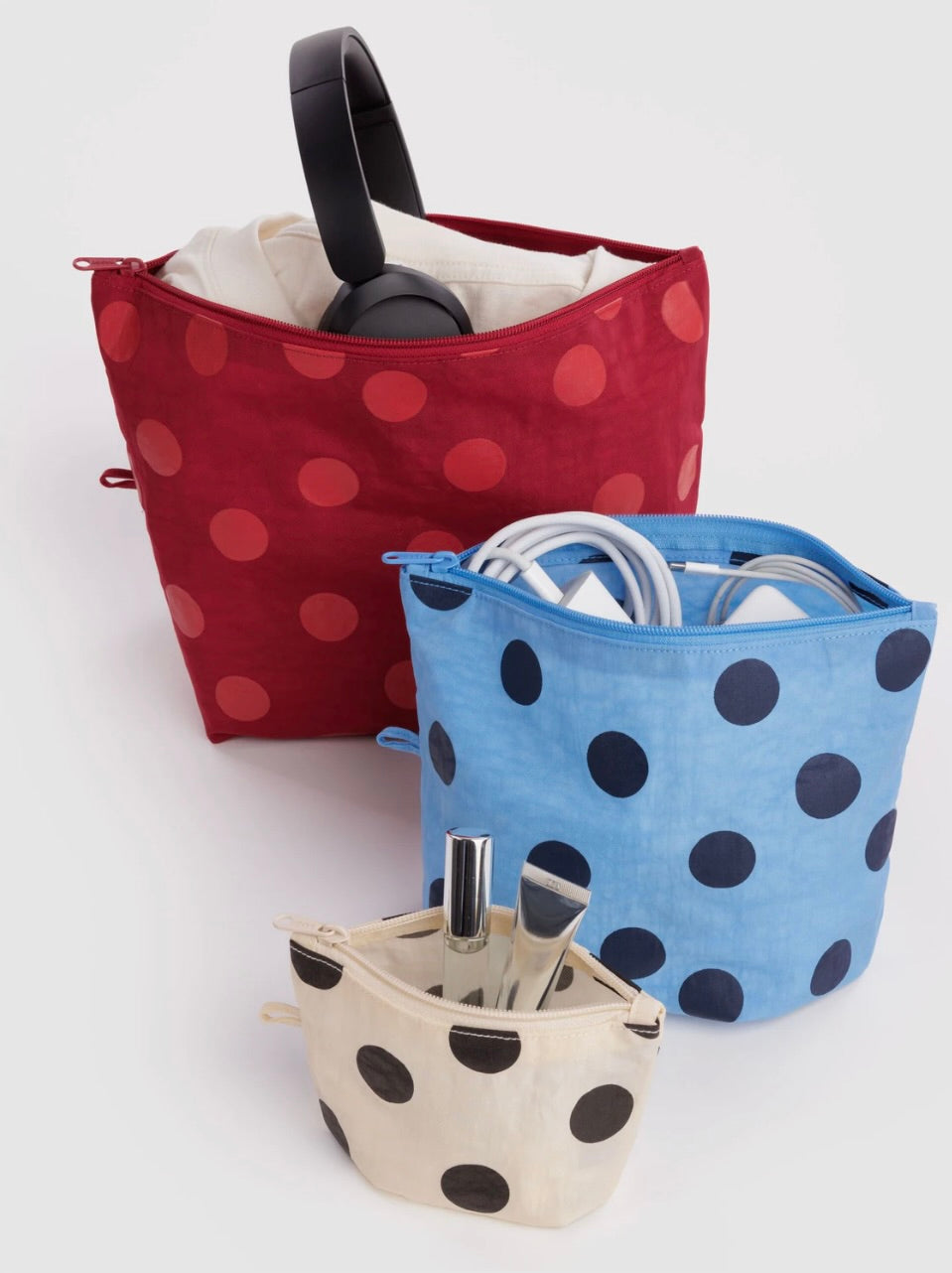 Three fabric zippered organizer pouches with polka dots, shown on a light gray studio background. The back pouch is dark red with red polka dots and contains black headphones; the middle pouch is light blue with dark blue polka dots and contains white cords; the front pouch is cream with black polka dots and contains a silver umbrella and pen.