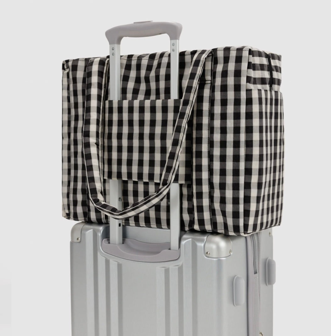 A black and white gingham checkered travel tote bag with a telescoping handle, shown resting on top of a silver hard-shell suitcase against a light gray background.