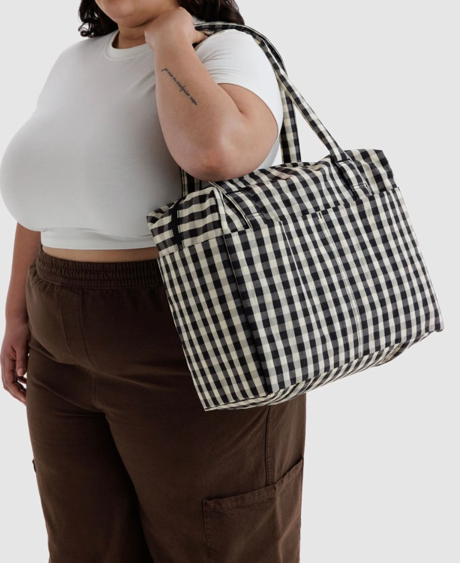 A person wearing a white crop top and brown pants carries a large black and white gingham check tote bag over their shoulder against a light gray background.