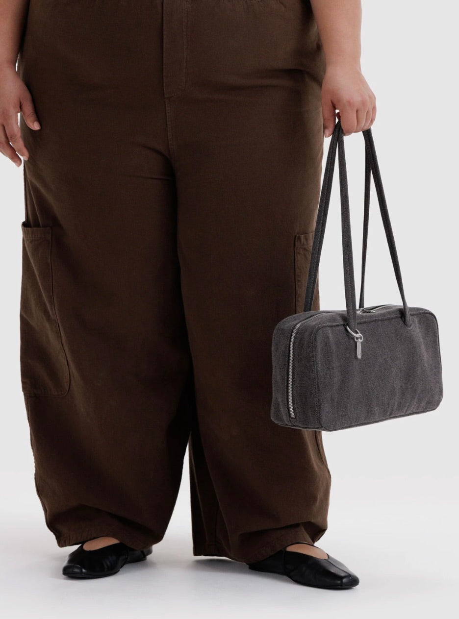 A person wearing loose brown cargo-style pants and black flats holds a dark gray rectangular shoulder bag with long handles against a light gray studio background.