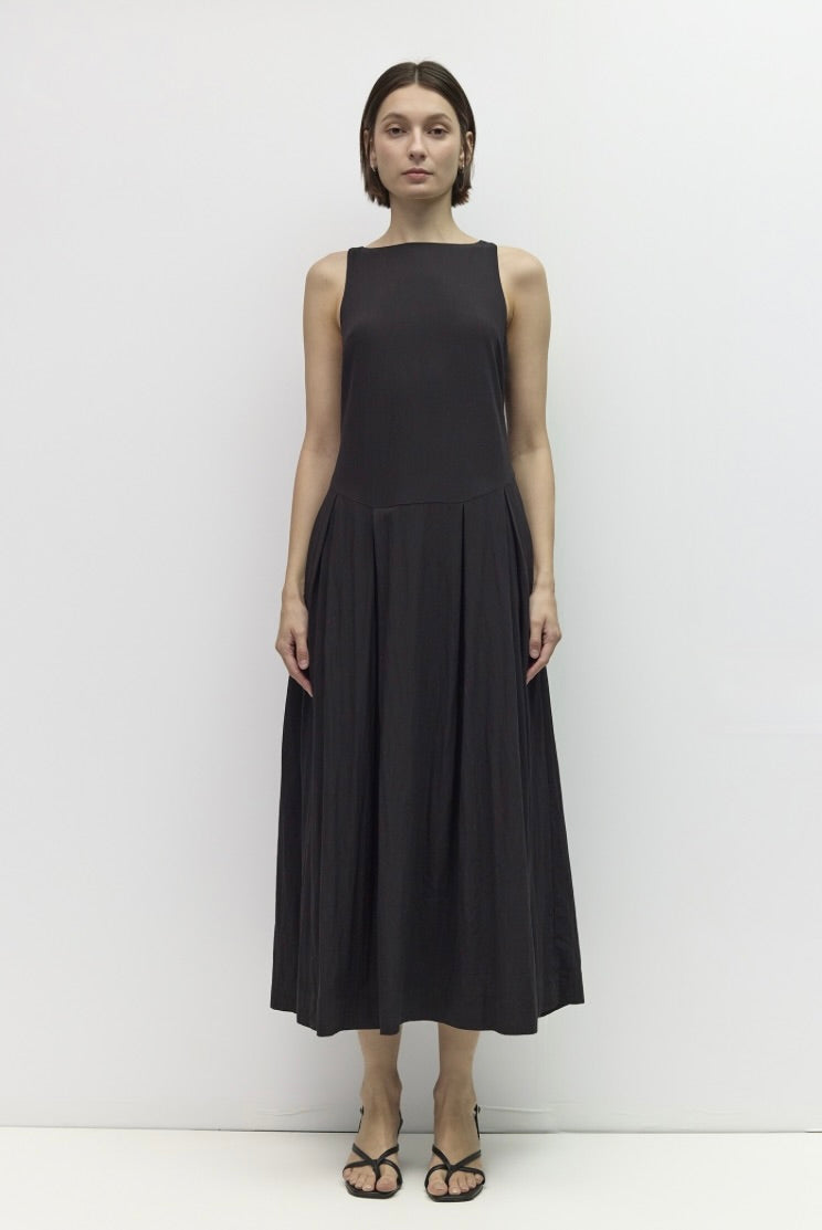 A woman stands against a plain white studio background wearing a sleeveless black midi dress with a structured bodice and flowing skirt, paired with black strappy heels.