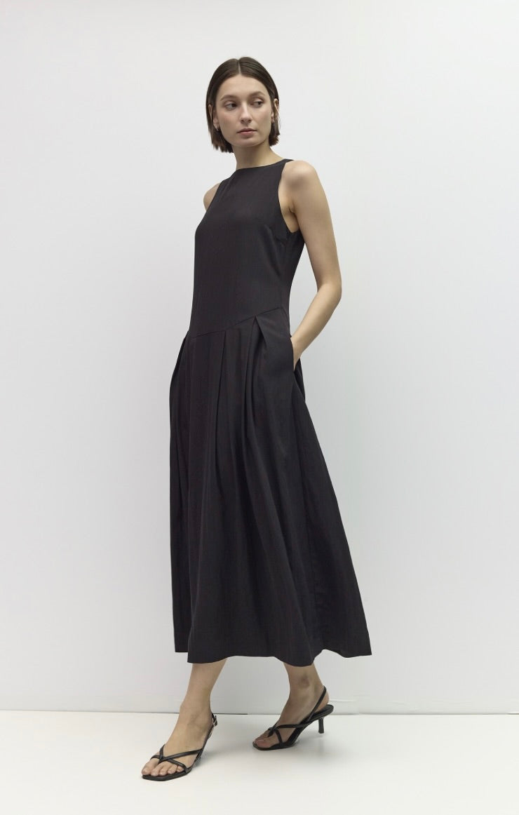 A woman modeling a sleeveless black midi dress with a high neckline and pleated skirt, paired with black strappy heels, against a plain white studio background.